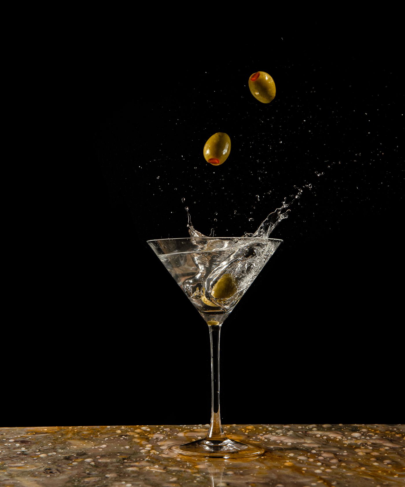 Martini glass with olives splashing in, against a black background